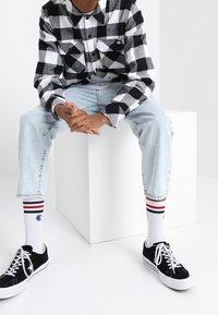 Svart och vit rutig flanellskjorta, ljusblå jeans, svarta sneakers och vita strumpor med röda och blå ränder, sittande på en vit kub.