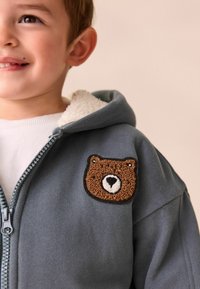 Ung liten flicka som ler, iklädd en grå zip-up hoodie med en texturerad brun björnfans på bröstet.