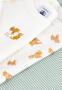 To hvite babyskjorter i bomull med gule leopardmønstre og teksten "Petit Bateau". Bakgrunnen har et grønt stripet stoff.