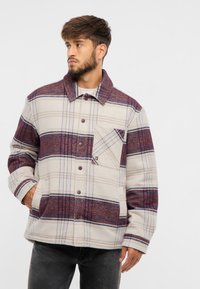 Jachetă din fleece bej cu un model în carouri maronii și albastre, având un închidere cu nasturi, guler și un buzunar pe piept.