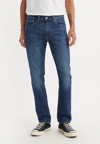 Homme portant un jean bleu droit, des baskets noires et blanches, et un t-shirt blanc, debout devant un fond clair uni.