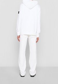 Vit hoodie med huva och framficka modell känguru, tillsammans med vida vita byxor, visad bakifrån.