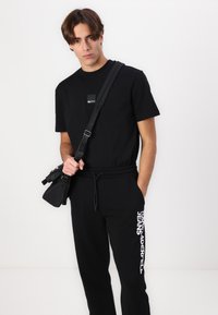 Melna īsām piedurknēm t-krekls ar mazu logo uzlīmi, apvienots ar melnajām sporta biksēm ar baltu zīmolu uz sāna. Ietver melnu crossbody somu.