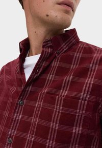 Rode geruite shirt gemaakt van textuurstof. Bevat een klassieke kraag, knoopdetails en een borstzak aan de linkerkant. Draagt over een wit shirt.