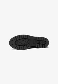 Black rubber shoe sole with deep treads and ridges, featuring a textured surface and a central logo embossed.