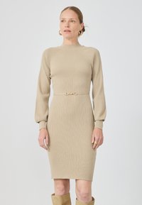 Beige ribbet strikkjole med lange ærmer, høj halsudskæring og guldtonet spænde detalje i taljen. Glat tekstur, tætsiddende design.