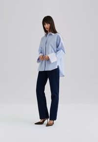 Ljust blårandig oversized skjorta med vit krage och manschetter, kombinerad med mörkblå raka jeans och bruna spetsiga klackar.