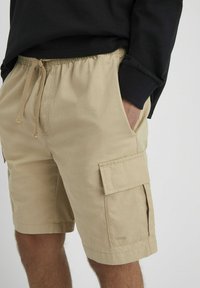 Shorts cargo beiges avec taille élastique et cordon de serrage. Comprend des poches latérales et une poche appliquée sur la cuisse. Texture en coton lisse.