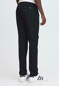 Homme portant un pantalon noir et des baskets blanches Adidas, vu de dos contre un fond clair.