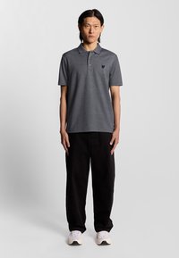 Mørkegrå polo skjorte med krave, korte ærmer og et lille sort logo på venstre bryst, parret med sorte bukser og hvide sneakers.