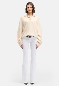 Maglione beige oversize con scollo a collo e chiusura con bottoni, abbinato a jeans bianchi a zampa e scarpe nere con tacco alto.