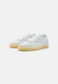 Sneaker bianchi realizzati in camoscio e pelle, caratterizzati da un design perforato, lacci e suole in gomma leggera color tortora. Presentano accenti blu sottili.
