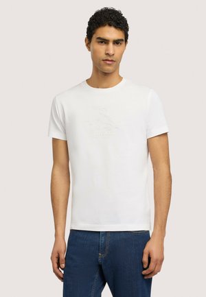 Młody mężczyzna z kręconymi włosami, ubrany w białą gładką koszulkę z krótkim rękawem i niebieskie dżinsy, na jasnym tle.