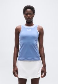 Γυναίκα που στέκεται με το σώμα στραμμένο προς τα εμπρός, φορά αμάνικο αθλητικό τοπ σε ανοιχτό γαλάζιο χρώμα με το λογότυπο «EA7» και λευκή φούστα, μπροστά σε απλό φόντο.