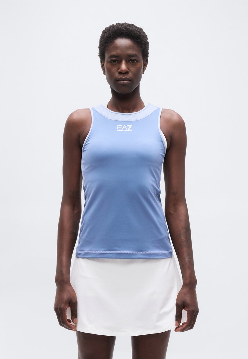 Γυναίκα που στέκεται με το σώμα στραμμένο προς τα εμπρός, φορά αμάνικο αθλητικό τοπ σε ανοιχτό γαλάζιο χρώμα με το λογότυπο «EA7» και λευκή φούστα, μπροστά σε απλό φόντο.