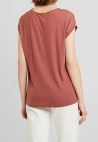 Osoba w różowej, krótkiej bluzce z podwiniętymi rękawami i jasnobeżowych spodniach, widoczna z tyłu na jednolitym szarym tle.