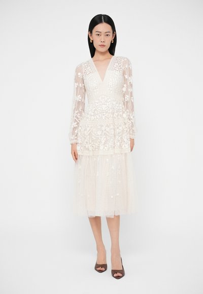 Femeie purtând o rochie midi albă din dantelă florală, cu mâneci lungi transparente și decolteu în V, asortată cu pantofi de culoare închisă, cu degetele descoperite, stând pe un fundal simplu.