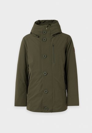 Homme portant une veste à capuche vert olive, un t-shirt blanc, un pantalon large marron et des baskets beiges, debout avec une main dans la poche.