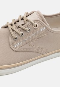 Beige kanvas sneakers med snørebånd, med en teksturert overdel, merket side detaljer og en gummisåle med et mønstrete mønster.