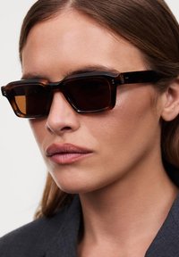 Lunettes de soleil écaille de tortue marron avec des montures épaisses et des verres foncés. Forme rectangulaire, texture lisse, avec de petites accents argentés.