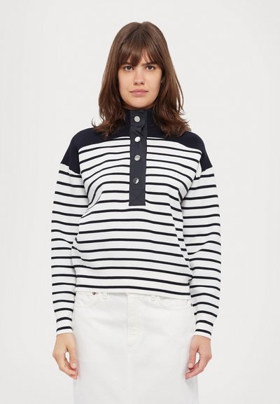 Stripete genser i marineblått og hvitt, med høy hals, knapplisting og lange ermer. Glatt tekstur og avslappet passform. Hvit skjørt nedenfor.