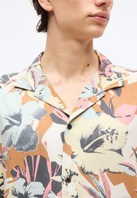 Νεαρό άτομο που φοράει ένα πουκάμισο με κουμπιά και εντυπωσιακά λουλούδια σε παστέλ αποχρώσεις, με μεγάλο γιακά σε χαλαρή γραμμή, γυρισμένο ελαφρώς προς τα δεξιά.