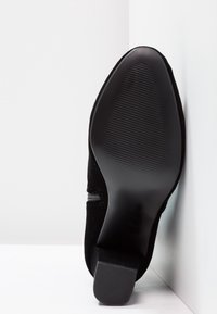 Chaussure en daim noir avec bout pointu, surface lisse et semelle en caoutchouc à motif avec un petit talon. Détail de fermeture éclair latérale visible.