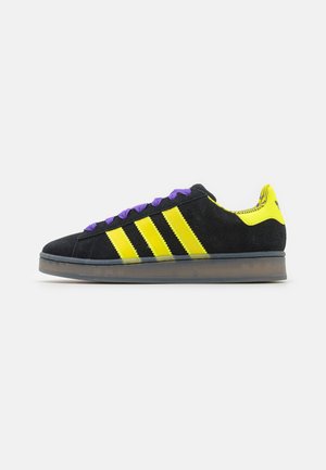 Baskets noires et jaunes avec trois bandes jaunes, lacets violets et semelle translucide, montrées de côté sur fond blanc.