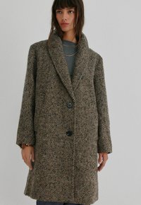 Brun houndstooth-mønstrede frakke lavet af tekstureret stof, med en bred sjalskrave, to knapper og sidelommer.