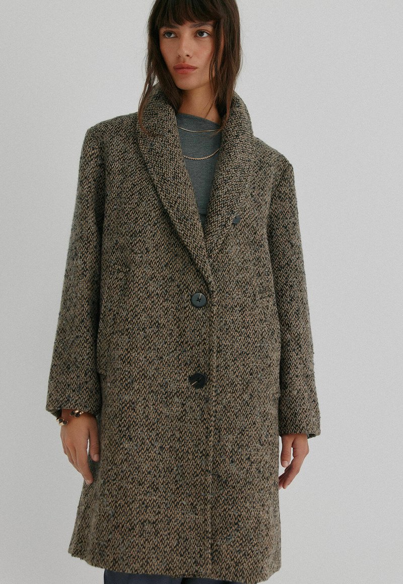 Brun houndstooth-mønstrede frakke lavet af tekstureret stof, med en bred sjalskrave, to knapper og sidelommer.