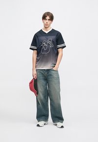 Μαύρη και γκρίζα διχτυωτή φανέλα με λευκό γιακά και ρίγες, με λογότυπο. Φοριέται με χαλαρές μπλε τζιν και κρατάει ένα κόκκινο καπέλο.