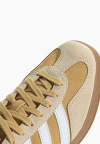 Rudi zomšiniai sportbačiai su baltais dryžiais, tekstūruota viršutinė dalis ir smėlio spalvos akcentai. Savybės: apvalus nosis, plokščios šnerves ir guminis padas.