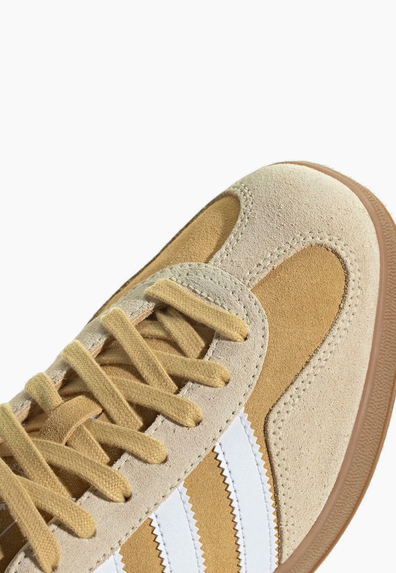 Braune Wildleder-Sneaker mit weißen Streifen, strukturiertem Obermaterial und beigen Akzenten. Merkmale sind eine runde Zehenform, flache Schnürsenkel und eine Gummisohle.