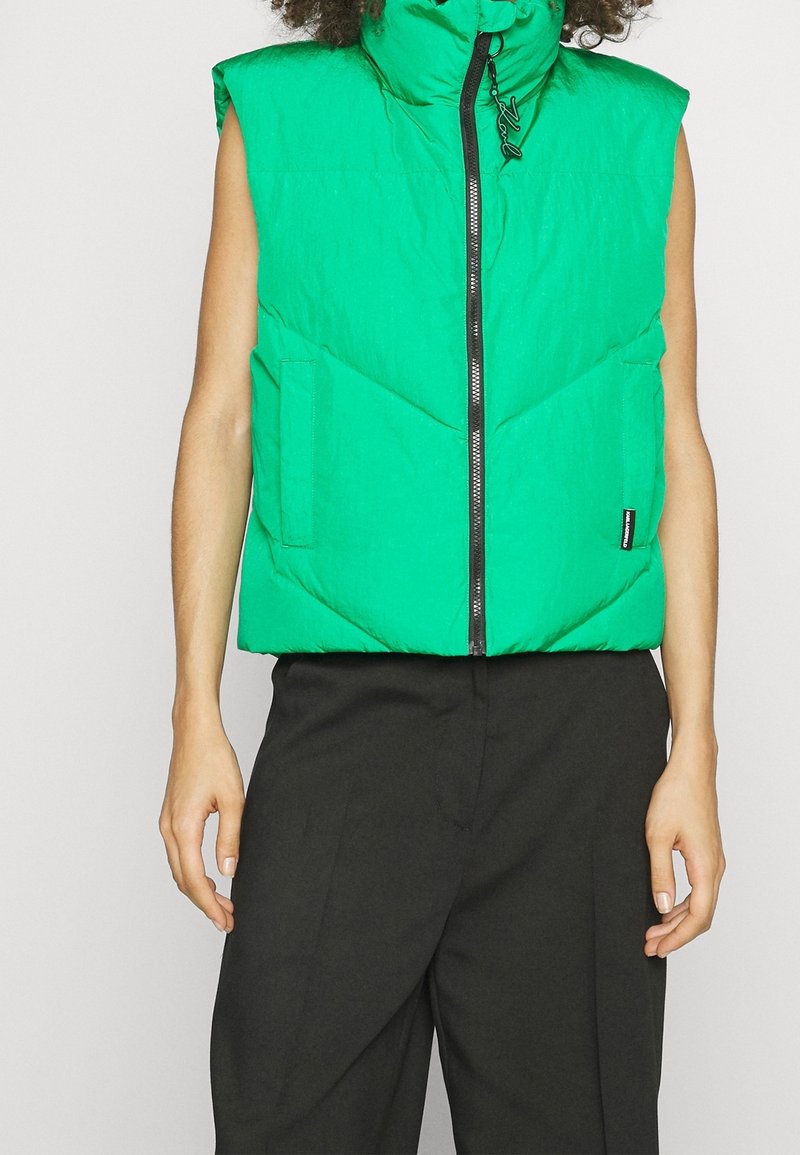 Groene gewatteerde bodywarmer met een hoge kraag en een ritssluiting aan de voorkant. Heeft visgraatstiksels en zijzakken. Draagt over zwarte broeken.