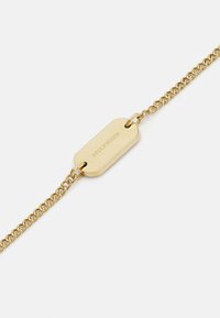 Pulsera de cadena tono dorado que presenta una etiqueta alargada con texto grabado, unida por pequeñas cadenas entrelazadas, con un acabado suave en toda la superficie.