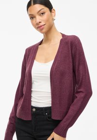 Burgundy cardigan with open front and long sleeves, made from soft texture. Features a ribbed hem and cuffs, worn over a white top and black jeans.