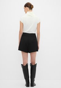 Top blanco de manga corta con escote drapeado, combinado con pantalones cortos negros de talle alto. La modelo lleva botas negras altas con un acabado texturizado.