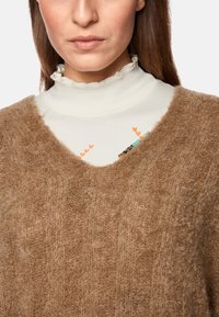 Brauner, strukturierter Strickpullover mit V-Ausschnitt, über einem cremefarbenen Oberteil mit Rüschenausschnitt und bunten grafischen Details getragen.
