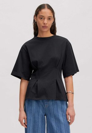 Vrouw met lang haar draagt een zwart topje met korte mouwen en plooitjes en een blauwe gestreepte broek met wijde pijpen, poseert voor een eenvoudige lichte achtergrond.