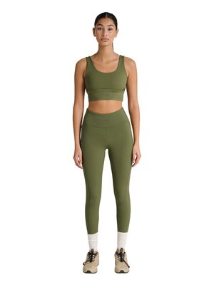 Kvinde står frontalt iført olivengrøn sports-bh og leggings med beige sneakers og hvide sokker mod hvid baggrund.