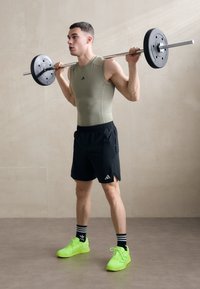 Athletic outfit featuring a fitted olive tank top, black shorts, and bright neon green shoes. Subject holds a barbell with weight plates.