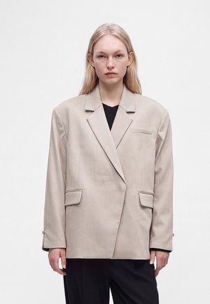 Blonde Frau mit glattem Haar trägt einen übergroßen beigen Blazer über einem schwarzen Top und steht vor einem schlichten hellen Hintergrund.