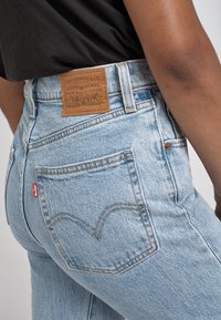 Personne portant un jean Levi's bleu clair avec une poche arrière visible, une étiquette rouge Levi's et une étiquette en cuir marron sur la ceinture, chemise noire partiellement rentrée.