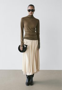 Bruine coltrui met lange mouwen, crèmekleurige geplooide maxi-rok, zwarte enkellaarsjes en een kleine zwarte handtas. Soepele stof en nauwsluitend ontwerp.