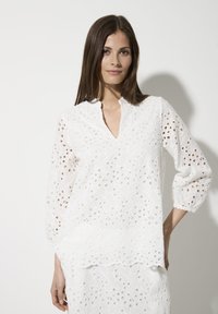 Weißes Blusenshirt mit V-Ausschnitt, 3/4-Ärmeln und filigranen Durchbruchdetails. Weicher, leichter Stoff mit fließender Silhouette und gerundetem Saum.
