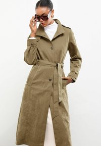 Vrouw in een beige trenchcoat met riem die oversized donkere zonnebril aanpast, gekleed in een witte turtleneck en gouden creolen, tegen een effen achtergrond.