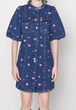 Robe en denim avec manches courtes bouffantes, fermeture à boutons sur le devant, poches poitrine, et motifs de cœurs roses sur tout le tissu.