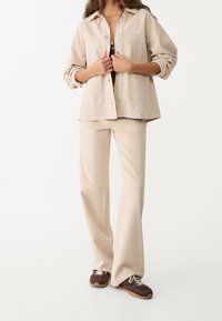 Beige oversized button-up jacket with chest pockets, paired with matching beige wide-leg pants and brown sneakers with light-colored laces.
