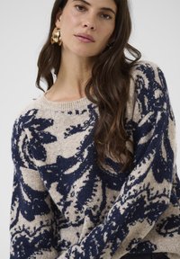 Žena s dlhými hnedými vlasmi, ktorá má na sebe veľké zlaté kruhové náušnice a béžový sveter s tmavomodrými abstraktnými kvetinovými vzormi.