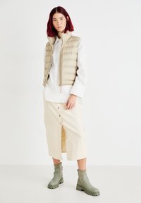 Chaleco acolchado beige con patrones cosidos, camisa blanca de botones, falda midi crema con una abertura frontal y botas de tobillo verde olivo con suelas gruesas.